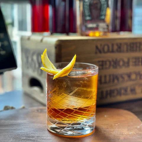 Cassia Burnt Sugar Manhattan Cocktail served in a crystal glass with a lemon twist, featuring a wooden bar backdrop.