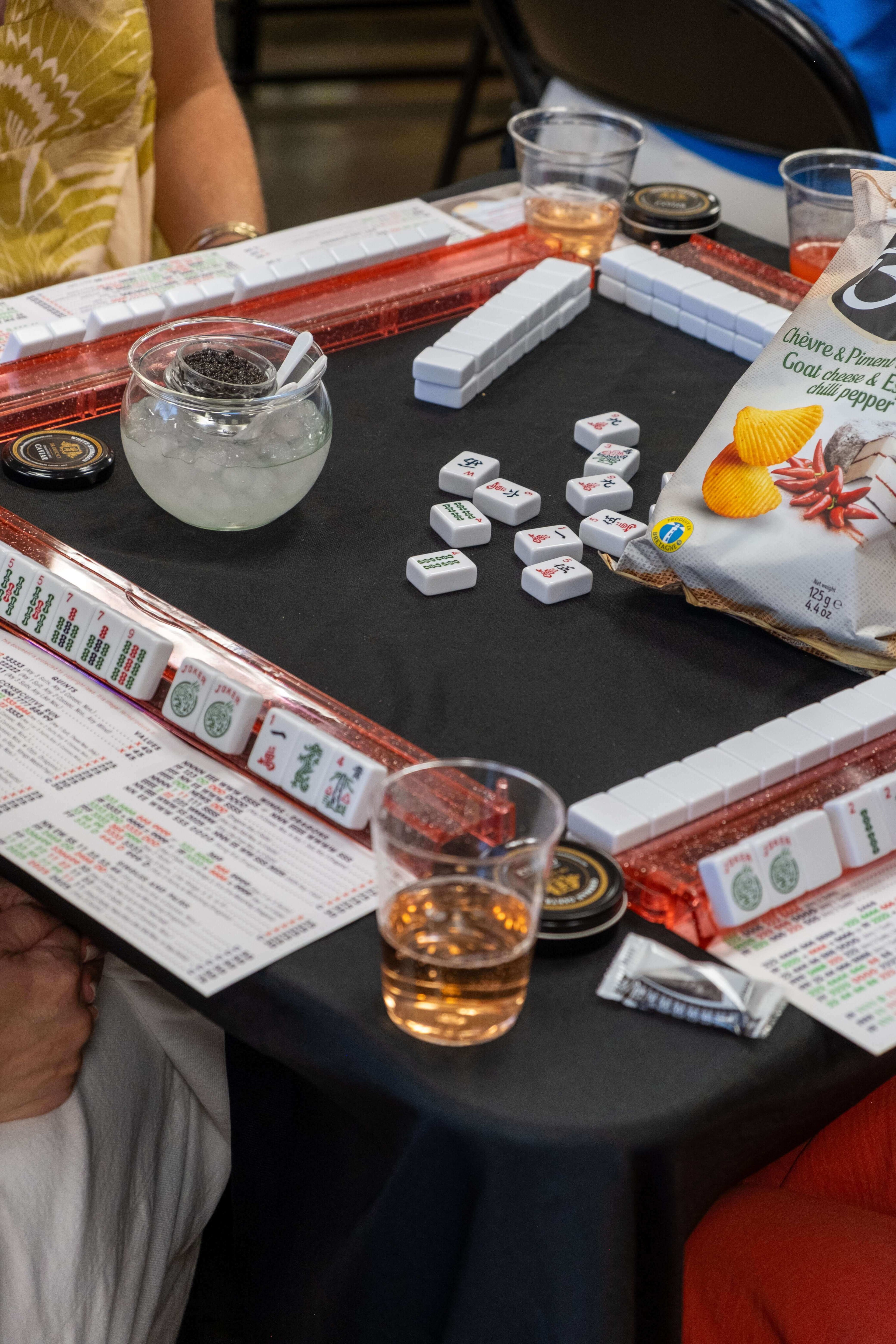 A close-up of a mahjong table with tiles, drinks, and snacks during a game night.
