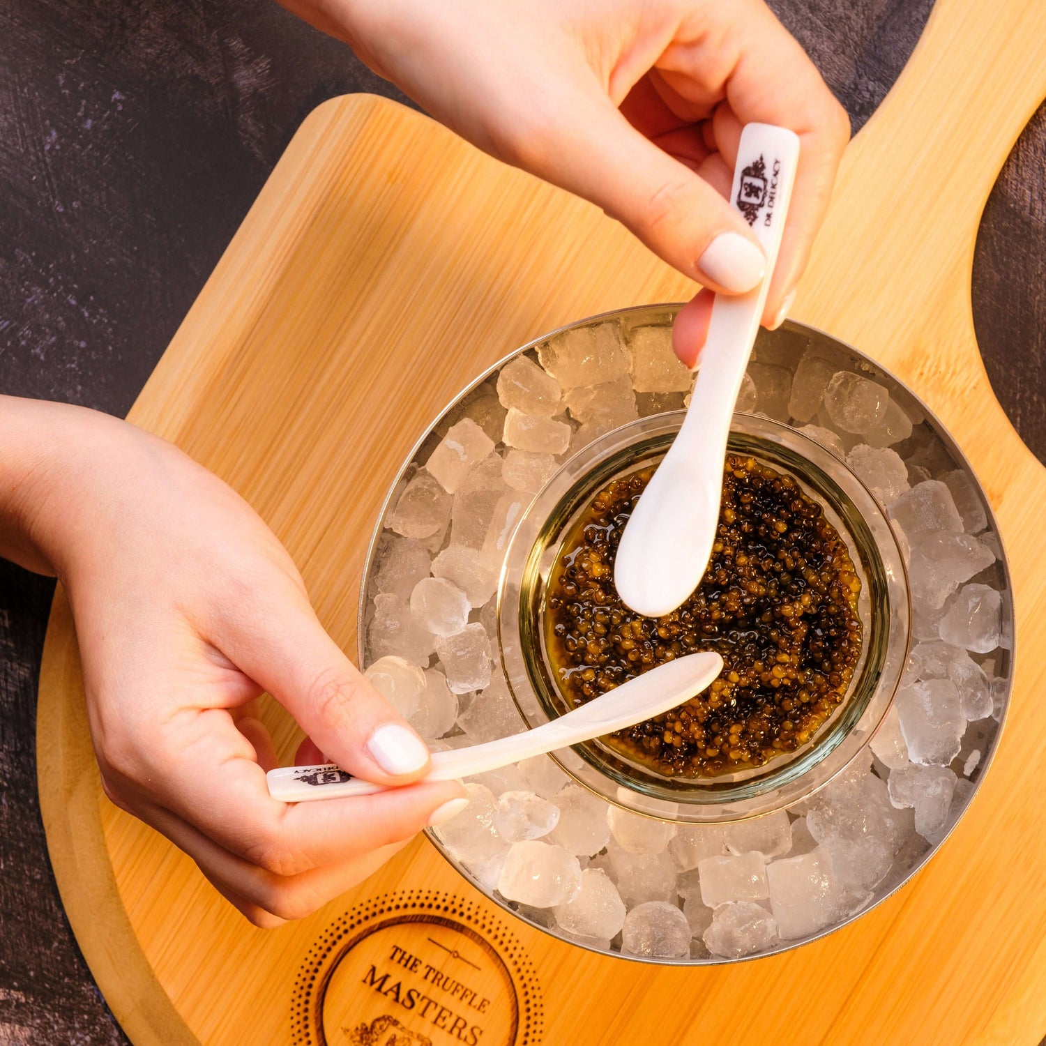 Hands using a Mother of Pearl Caviar Spoon to serve caviar in an elegant dish over ice.